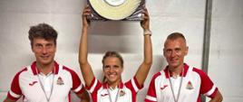 Widok na trzech strażaków w strojach sportowych, w biało-czerwonych koszulkach. W środku kobieta strażak, w rękach, nad głową trzyma talerz jako trofeum zawodów.