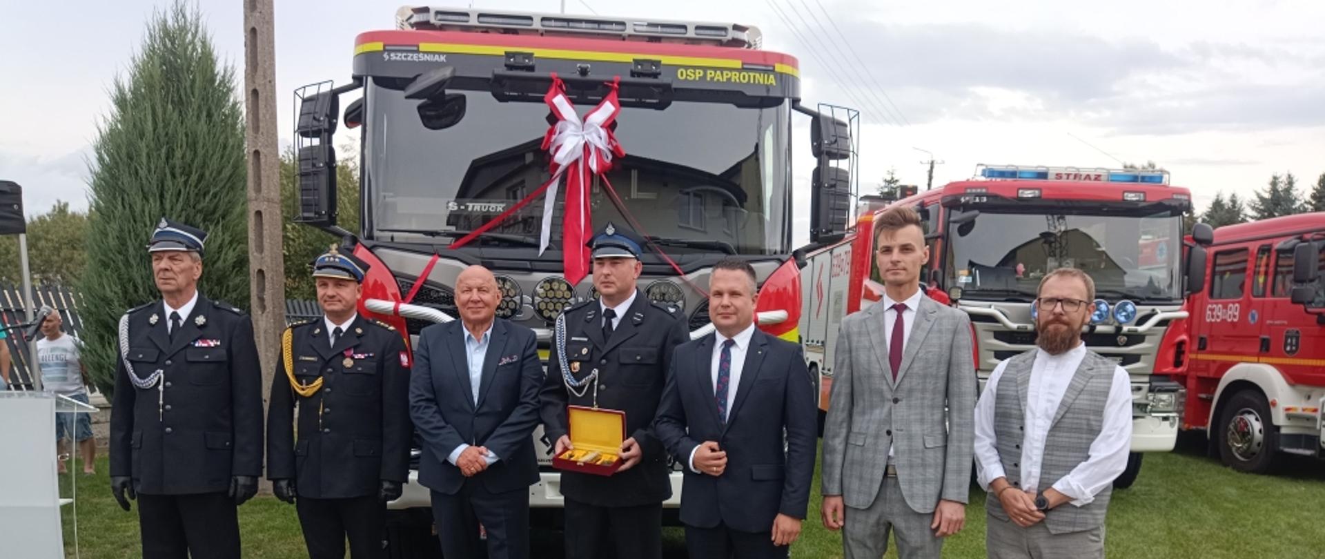 Na tym zdjęciu grupa mężczyzn, ubranych zarówno w mundury, jak i garnitury, pozuje do wspólnego zdjęcia. Stoją przed nowym, czerwonym wozem strażackim. Mężczyzna w garniturze trzyma w rękach złotą plakietkę w drewnianym pudełku, a w tle widać inny wóz strażacki.