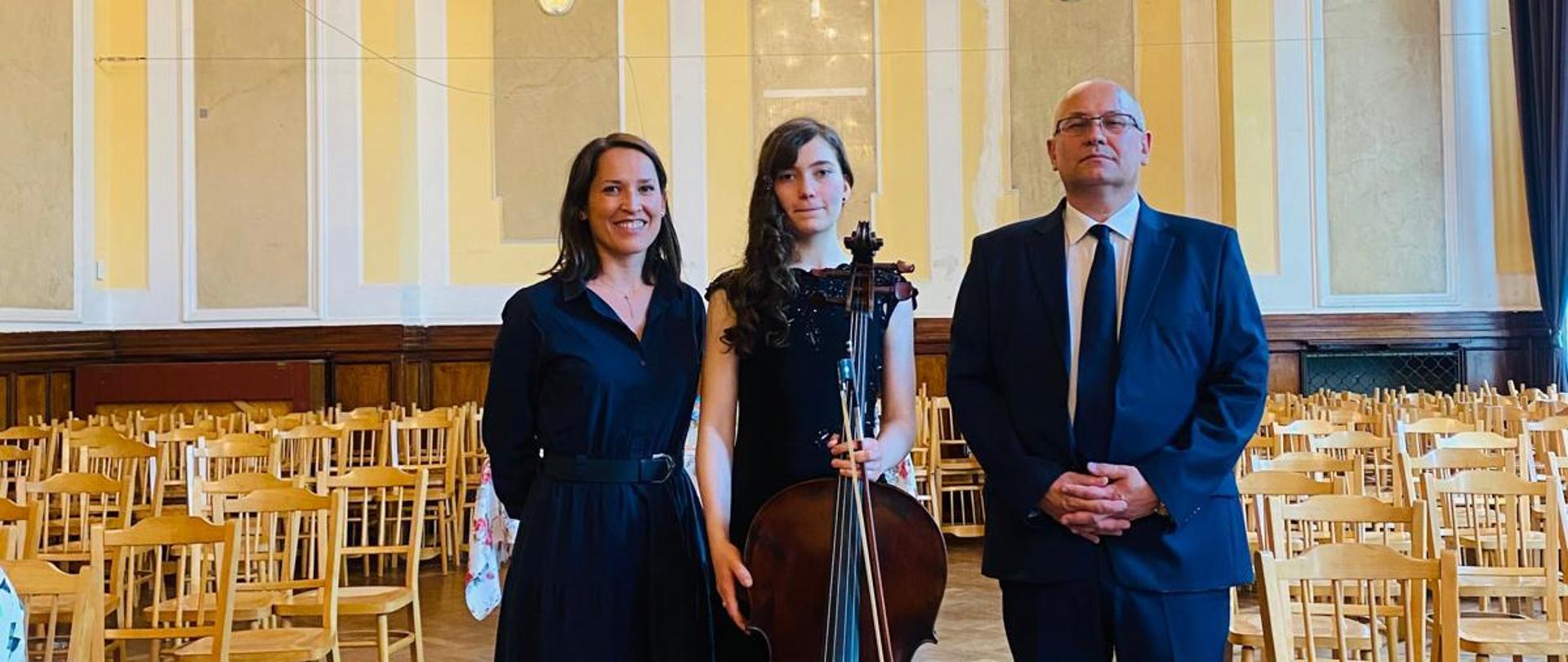 Zdjęcie przedstawia wiolonczelistkę która stoi trzymając w ręku smyczek i wiolonczelę po jej lewej stronie stoi pani profesor a po prawej stronie pan akompaniator w tle widoczne krzesła oraz żółto białe ściany i parkiet