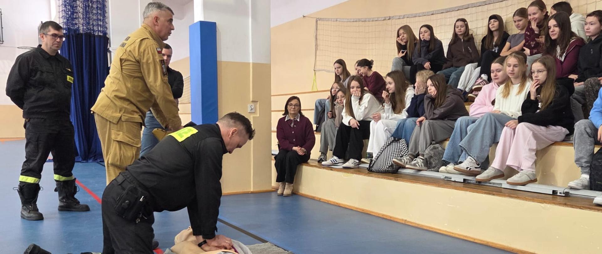Strażacy przeprowadzają pokaz pierwszej pomocy przedmedycznej dla grupy młodzieży zgromadzonej na trybunach sali gimnastycznej. Jeden z ratowników demonstruje prawidłową technikę wykonywania masażu serca na fantomie medycznym leżącym na macie.