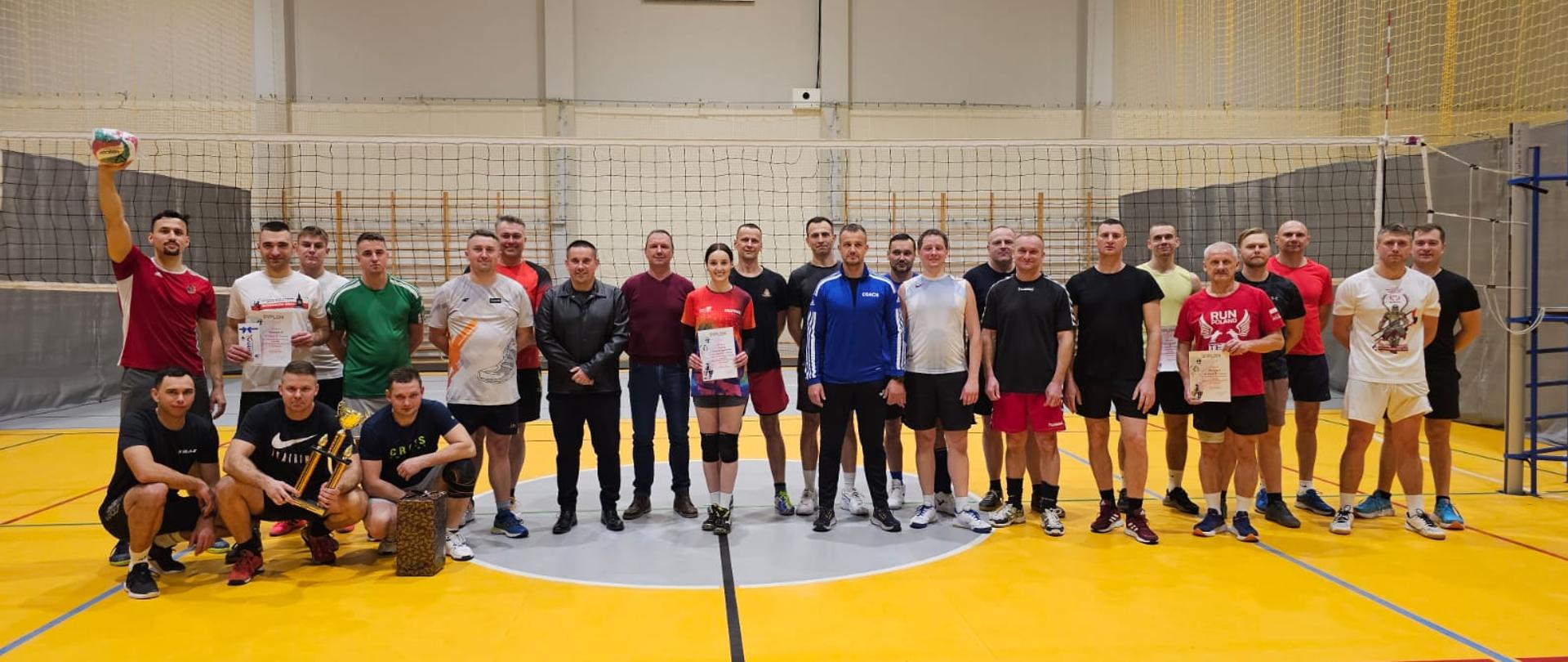 Zdjęcie grupowe na tle siatki przedstawia zawodników biorących udział w turnieju oraz komendanta powiatowego i prezesa CSIR. Zawodnicy trzymają w rękach nagrody rzeczowe, puchary oraz dyplomy. 