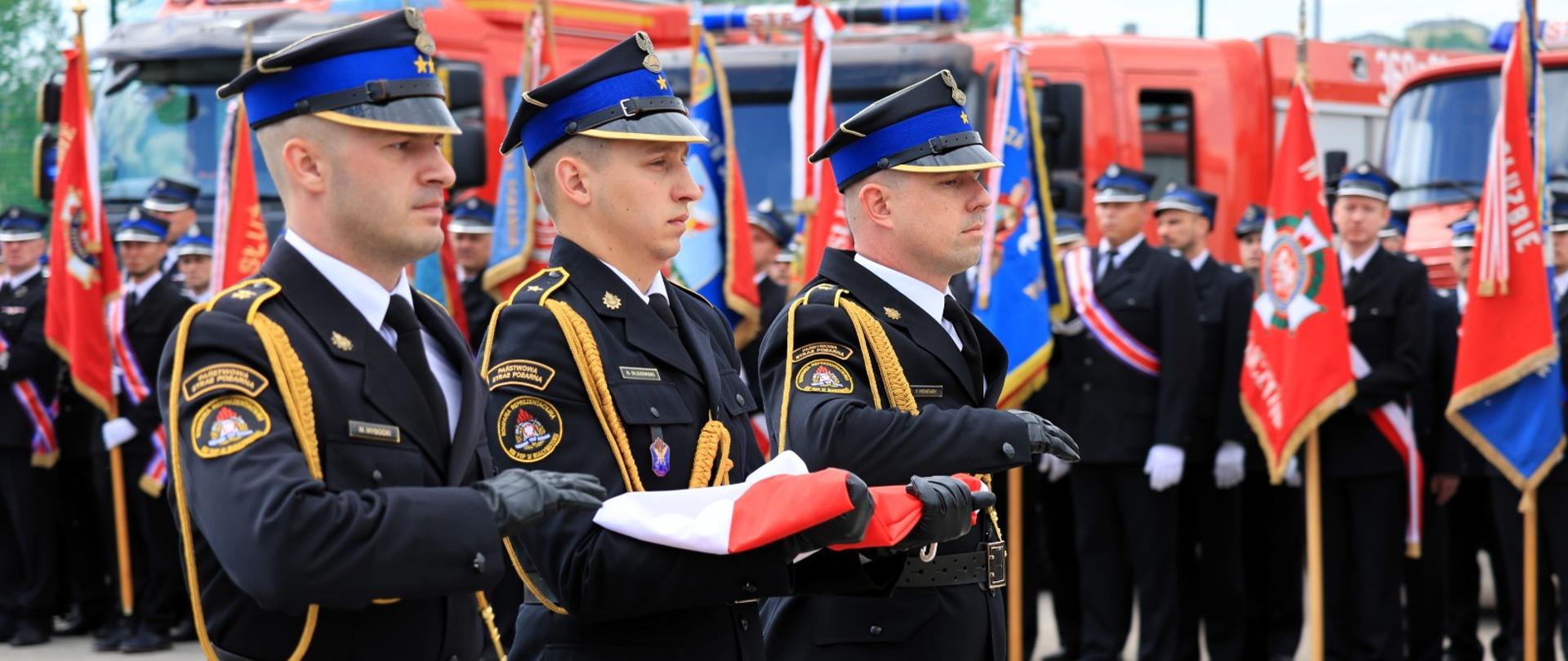 maszerujący poczet flagowy, w kierunku mocowania flagi. W tle widoczne pododdziały oraz samochody ratowniczo-gaśniczego 