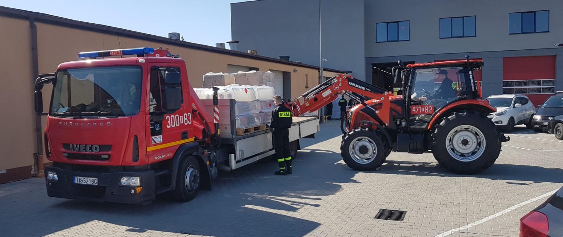 Na zdjęciu widać ciężarowy samochód strażacki ze skrzynią załadunkową na który strażak przy pomocy ciągnika z ładowaczem czołowym umieszcza na samochodzie palety z płynem do dezynfekcji