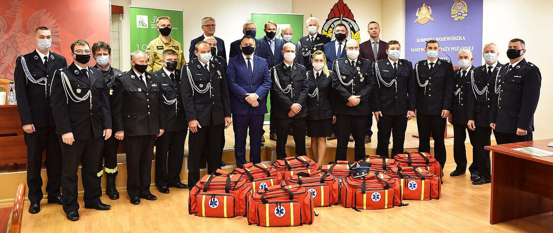 na zdjęciu grupa osób stojących w półkolu w pomieszczeniu, w skład grupy wchodzą strażacy OSP ubrani w mundury wyjściowe, strażak PSP w umundurowaniu służbowym w kolorze piaskowym oraz kilku mężczyzn ubranych w garnitury, przed zgromadzonymi na podłodze leżą torby - zestawy ratunkowe w kolorze czerwonym. Za zgromadzonymi ustawione kolorowe roll-upy