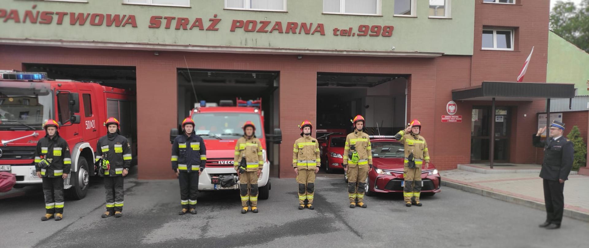 Strażacy stoją na baczność przed komendą, po prawej stronie stoi Komendant Powiatowy salutuje. Samochody strażackie wyprowadzone są z garażu