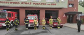 Strażacy stoją na baczność przed komendą, po prawej stronie stoi Komendant Powiatowy salutuje. Samochody strażackie wyprowadzone są z garażu