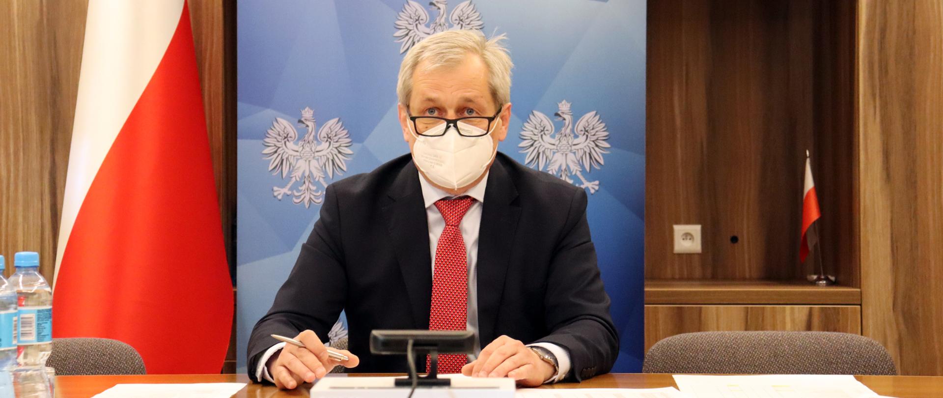 Na zdjęciu zasiadający za stołem wojewoda opolski Sławomir Kłosowski. Dłonie trzyma nad kartką, która leży przed nim na stole. Jedną dłoń ma uniesioną, w której trzyma długopis. Za wojewodą widać roll up z logo Wojewody Opolskiego oraz biało-czerwoną flagę, stojącą w pionie. Po lewej stronie kadru na dole widać plastikowe butelki z wodą.