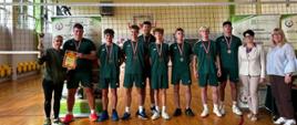 W hali sportowej, przed siatką do piłki siatkowej, stoi grupa zawodników ubranych w jednakowe ciemnozielone stroje sportowe. Chłopcy mają na szyjach medale. Po lewej stronie grupy stoi trenerka trzymająca w górze puchar, a jeden z zawodników prezentuje dyplom. Po prawej stronie stoją dwie kobiety. W tle widoczne są banery związane ze szkołami rolniczymi i organizowanymi zawodami.
