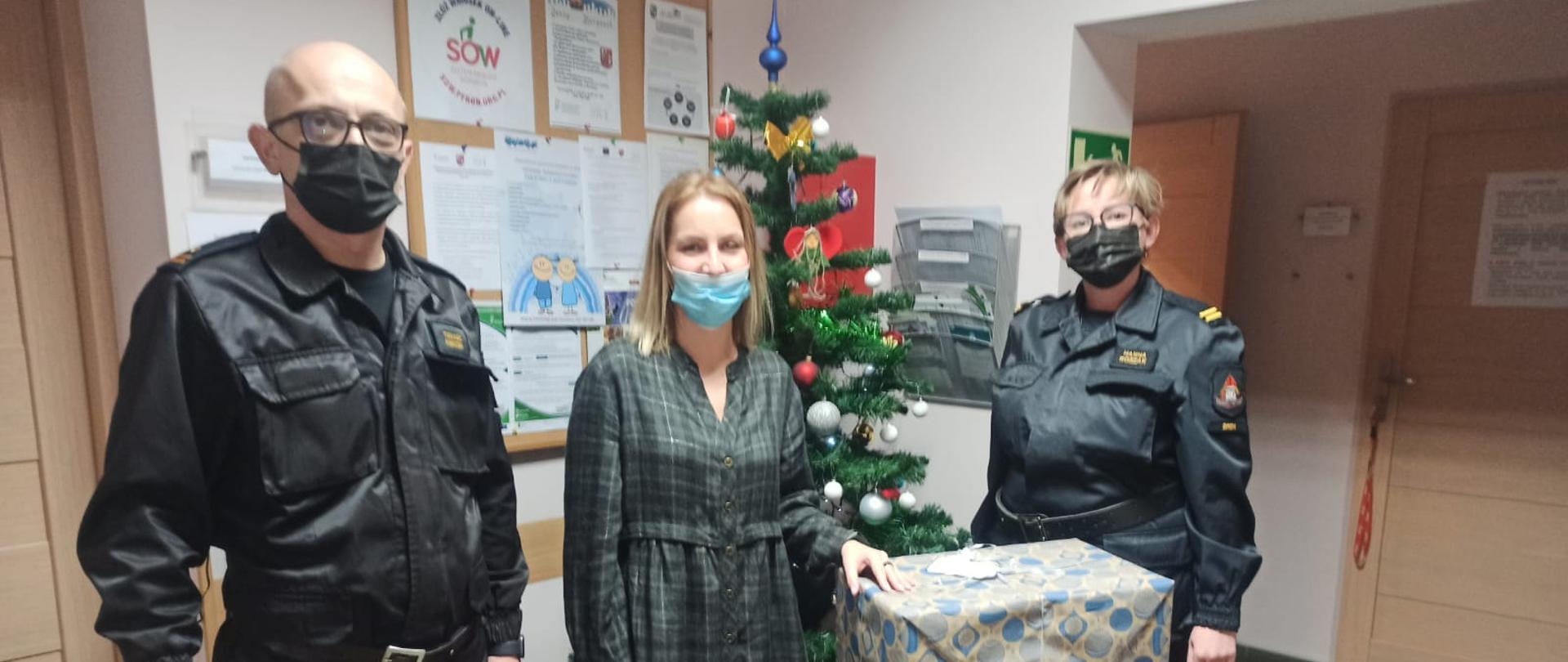 Na zdjęciu pracownik PCPR w Śremie, oraz dwóch funkcjonariuszy na tle choinki przed nimi zapakowane prezenty dla dzieci