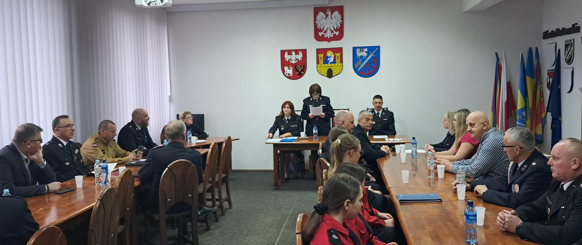 Sala posiedzeń. Z prawej lewej i na wprost stoły, za stołami siedzą strażacy oraz zaproszeni gości. Trwa zebranie.