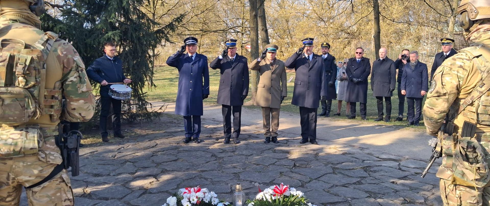 Czworo funkcjonariuszy różnych służb mundurowych w mundurach galowych stoi na kamiennej alejce i oddaje honor, salutując w kierunku miejsca pamięci. Naprzeciwko nich stoją żołnierze w pełnym wyposażeniu wojskowym, pełniący wartę honorową. W tle zgromadzeni są inni uczestnicy uroczystości oraz muzyk z werblem. Przestrzeń otaczają drzewa