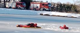 Kolorowa fotografia w słoneczny dzień wykonana na zalodzonym akwenie wodnym. Przedstawia ćwiczących dwóch strażaków ubranych w specjalne ubrania, jeden z nich znajduje się wodzie w wykonanym przeręblu, natomiast drugi odpowiednio zabezpieczony próbuje go wyciągnąć z wody.