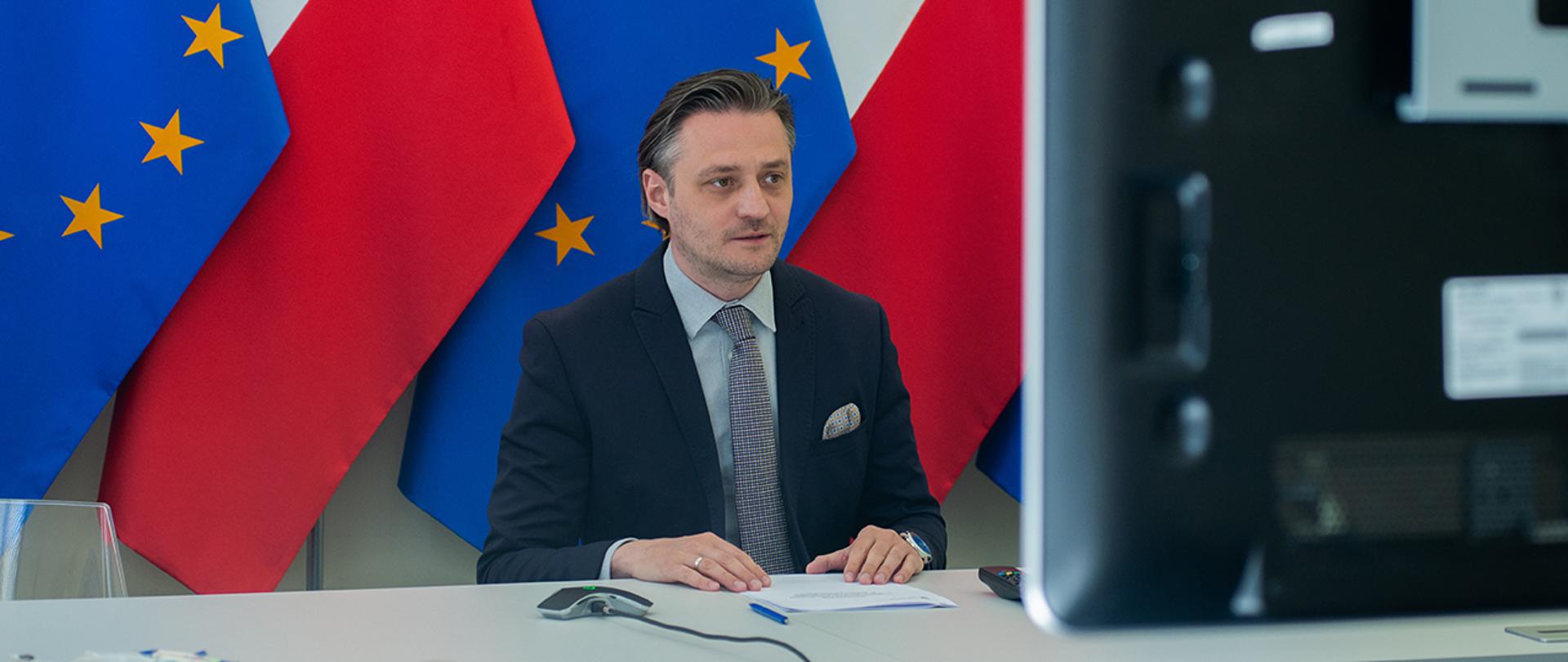 Na zdjęciu: wiceminister Bartosz Grodecki podczas wideokonferencji. W tle flagi Polski oraz UE