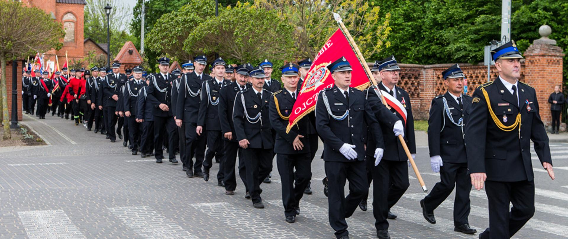 Ulicą Bukowca maszeruje pododdział druhów Ochotniczej Straży Pożarnej na czele idzie dowódca oraz poczet sztandarowy. W tle widać Młodzieżową Drużynę Pożarniczą oraz pozostałe poczty sztandarowe. 