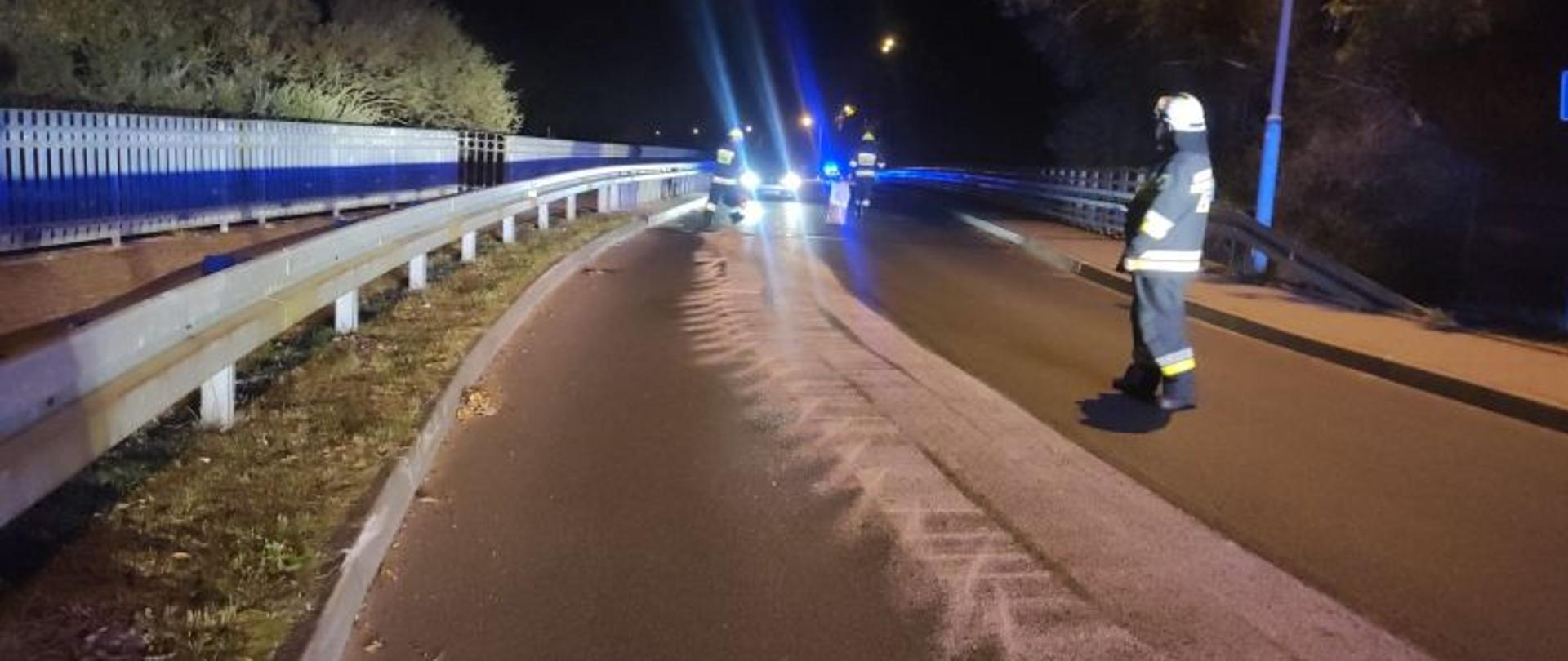 Zdjęcie wykonane w nocy. W tle ciemne, czarne niebo. Na bokach drogi barierki. Na środku jednego z pasów ślad od rozsypanego sorbentu. Przy śladzie widać stojącego strażaka. Zdjęcie rozświetlają lampy błyskowe.