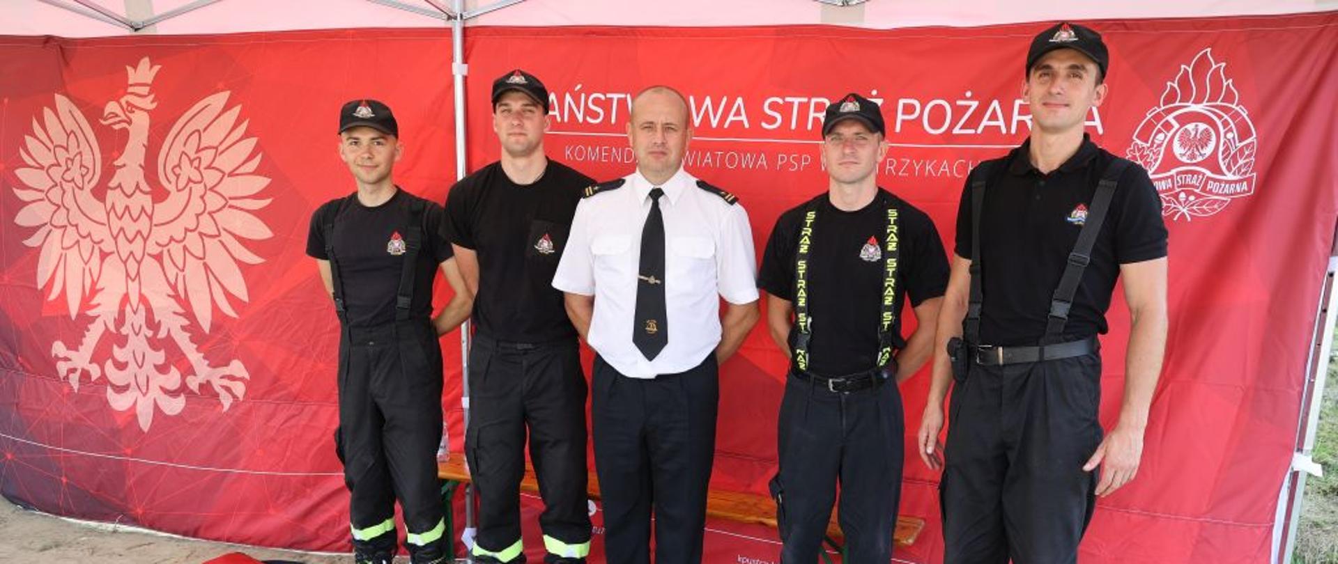 Na zdjęciu widać komendanta powiatowego PSP w Ustrzykach Dolnych oraz czterech funkcjonariuszy komendy powiatowej PSP w Ustrzykach Dolnych a także torbę medyczną. Za strażakami widać ścianę namiotu na której widnieje Godło Polski Orzeł Biały a także napisy Państwowa Straż Pożarna Komenda Powiatowa PSP w Ustrzykach Dolnych