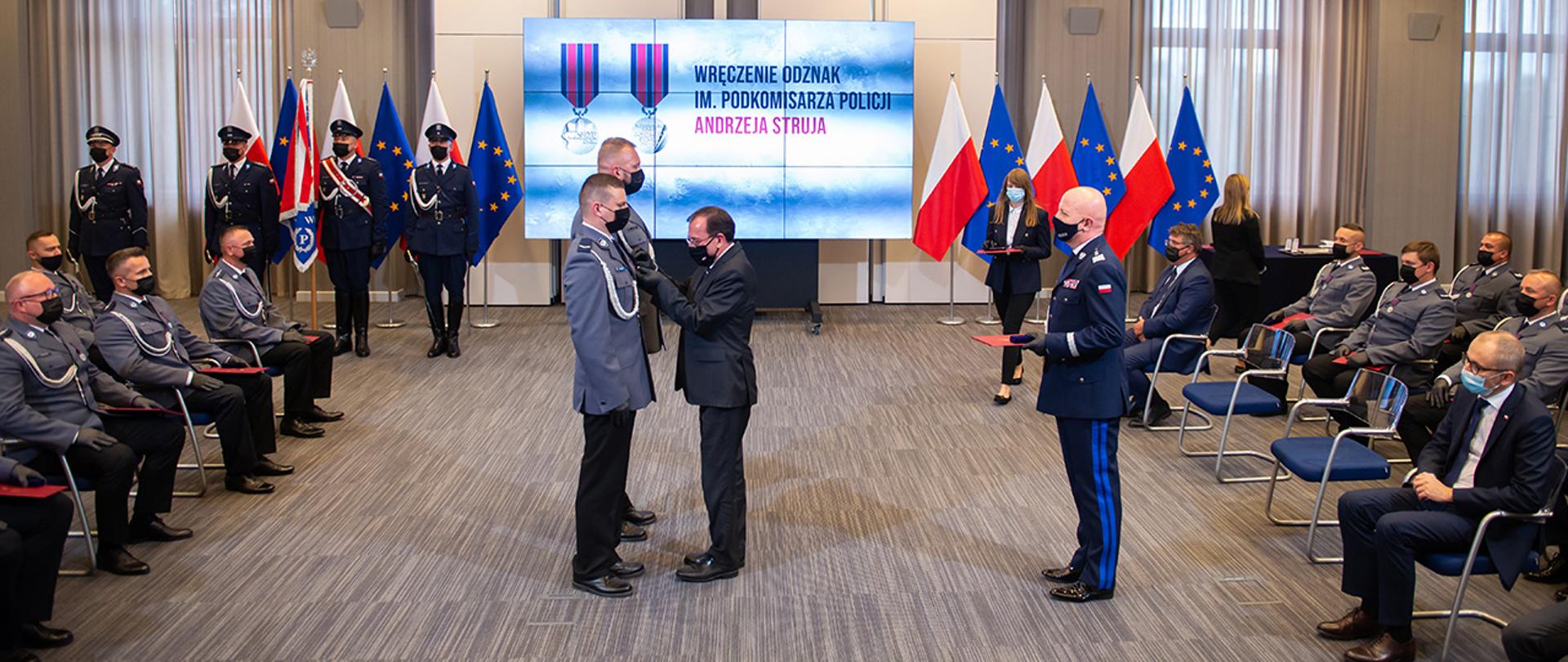 Minister Mariusz Kamiński przypina odznaki policjantom. Z tyłu stoi Komendant Główny Policji.