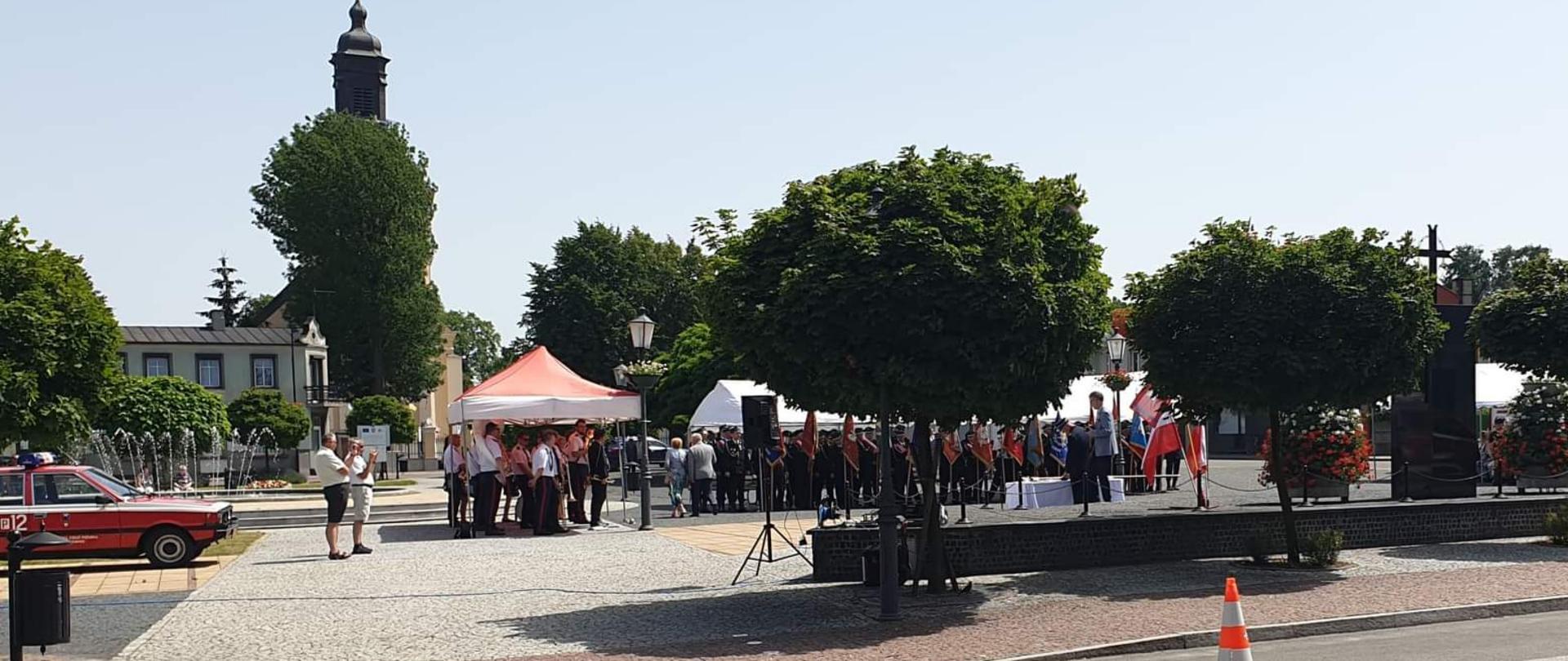 Na zdjęciu rynek w Koźminku. Po prawej pomnik. W tle budynek kościoła parafialnego. Niebo błękitne. Pogoda słoneczna. W centralnym miejscu widać osoby w mundurach i sztandary.