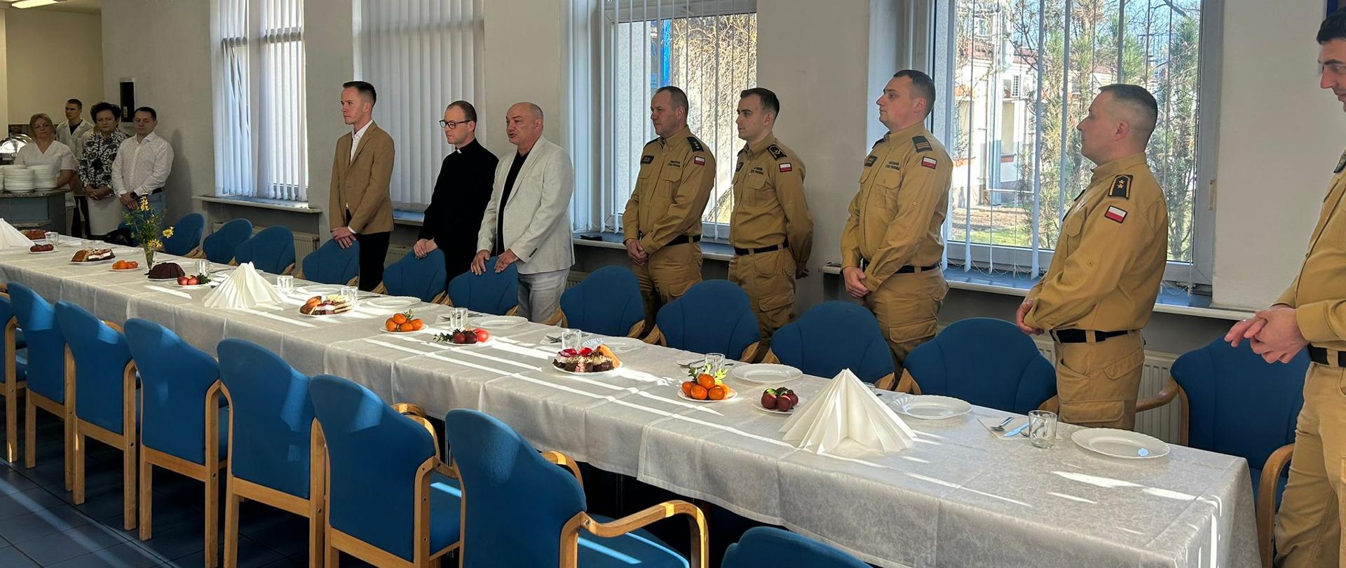 Przy długim stole ustawionym pod oknami stoją funkcjonariusze PSP w mundurach oraz kilka osób cywilnych i duchowny. Na stole znajdują się świąteczne potrawy i dekoracje wielkanocne. Przez duże okna wpada światło dzienne, oświetlając uczestników stojących w skupieniu podczas modlitwy