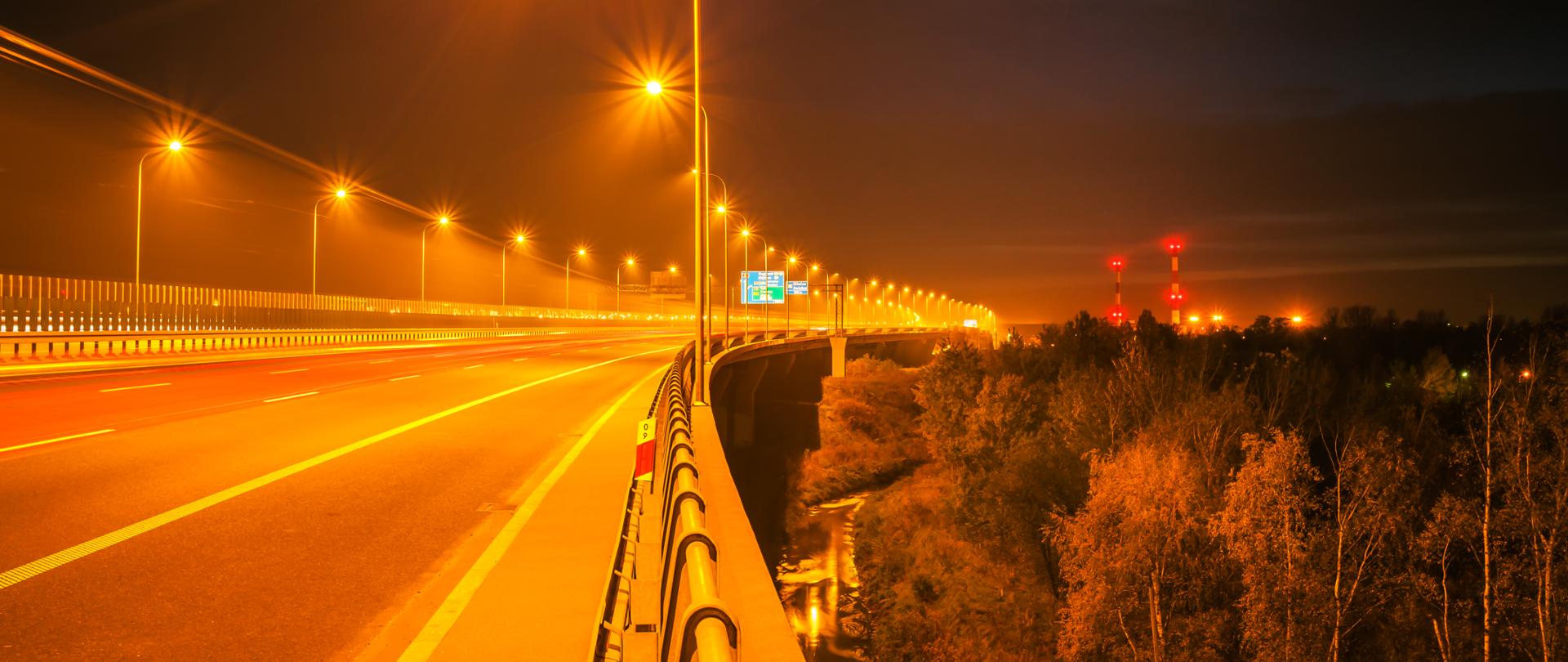 Fotografia nocna estakady w ciągu autostrady A1 w Maciejowie. Widoczne liczne latarnie drogowe oświetlające jezdnię estakady. Po prawej stronie widoczne na horyzoncie dwa wysokie kominy oświetlone czerwonym światłem. 