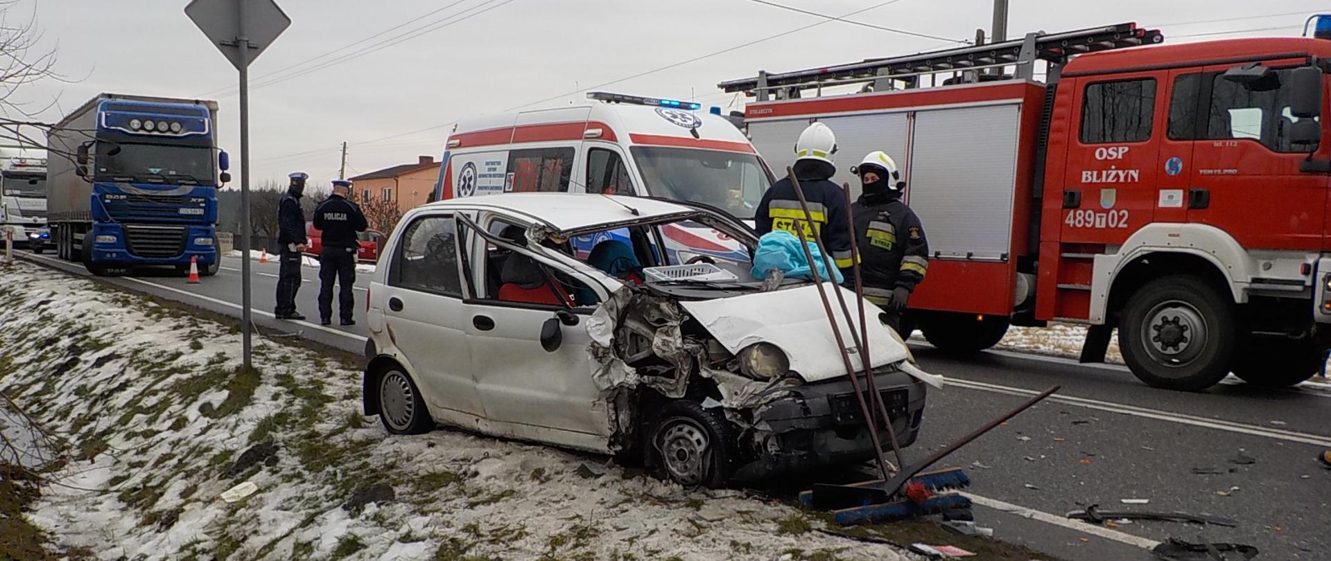 Na zdjęciu widać uszkodzony pojazd marki Daewoo Matiz znajdujący się na poboczu drogi, samochód gaśniczy jednostki OSP oraz karetkę pogotowia. Dwóch ratowników straży pożarnej usuwa skutki zdarzenia. Dwóch funkcjonariuszy Policji prowadzi czynności wyjaśniające zdarzenie.
