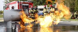 Na pierwszym planie palący się zbiornik na gaz LPG oraz opary, w głębi strażacy w umundurowaniu bojowym oraz samochód gaśniczy