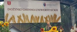 Na zdjęciu znajduje się scena plenerowa udekorowana z okazji dożynek, czyli tradycyjnego święta plonów. Nad sceną wisi biały baner z brązowym napisem „DOŻYNKI GMINNO-UNIWERSYTECKIE” oraz z grafiką przedstawiającą kłosy zboża. Po lewej stronie banera widnieje herb z białym krzyżem, po prawej — logo Uniwersytetu Rolniczego im. Hugona Kołłątaja w Krakowie. Na scenie, na tle zielonych drzew, stoi z mikrofonem pani Dyrektor Kasy Rolniczego Ubezpieczenia Społecznego w Krakowie Iwona Szablińska. Przed nią ustawiono dużą, białą donicę z pięknie rozkwitłymi słonecznikami. Na scenie znajdują się też inne osoby, w tym kobieta w jasnej sukni oraz mężczyzna ubrany w ciemny garnitur. Scena ozdobiona jest dodatkowo dyniami i koszami wiklinowymi, co podkreśla jesienny i rolniczy charakter wydarzenia. Całe wydarzenie odbywa się na świeżym powietrzu.