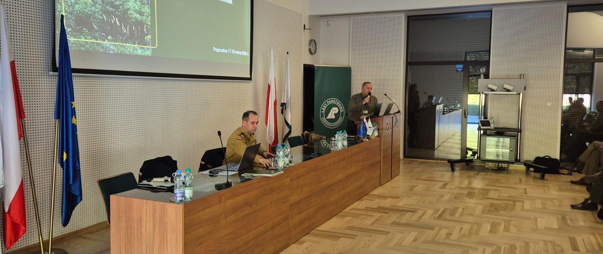 Zachodniopomorski komendant wojewódzki PSP podczas prezentacji o ochronie przeciwpożarowej w lasach. komendant siedzi przy stole. Za jego plecami znajduje się ekran projekcyjny na którym wyświetlana jest prezentacja. Zdjęcie powiększy się po kliknięciu na nie.