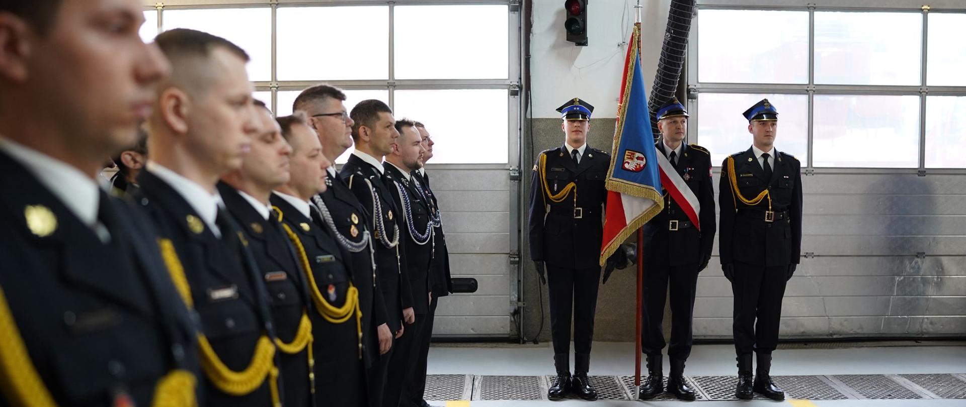 Na fotografii widać po lewej stronie strażaków stojących w rzędach, ubranych w granatowy mundur przepasany złotym i srebrnym sznurem, na wprost stoi poczet sztandarowy złożony z 3 osób również ubranych w mundury.