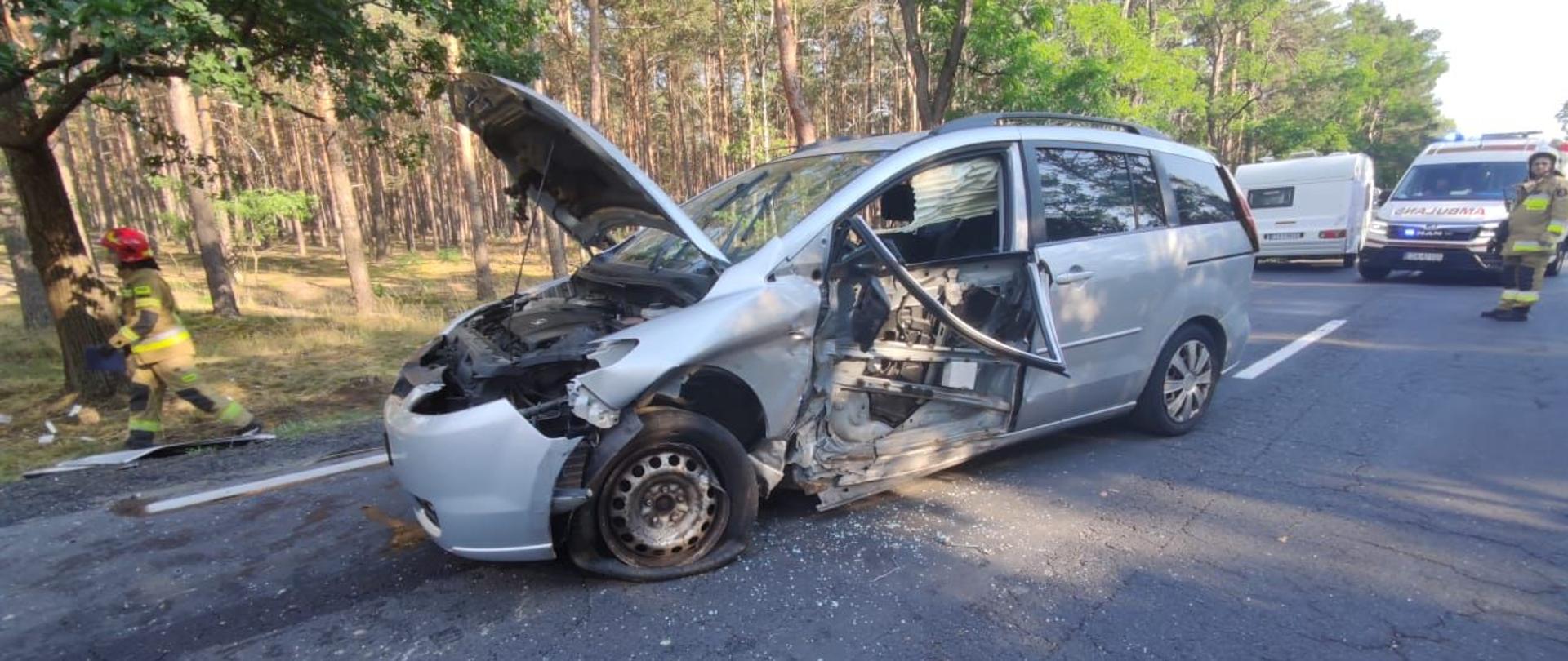 Na zdjęciu drogi krajowej numer dwadzieścia siedem stoi samochód osobowy marki Mazda pięć z rozwalonym bokiem pojazdu od strony kierowcy. Trwają działania ratownicze.