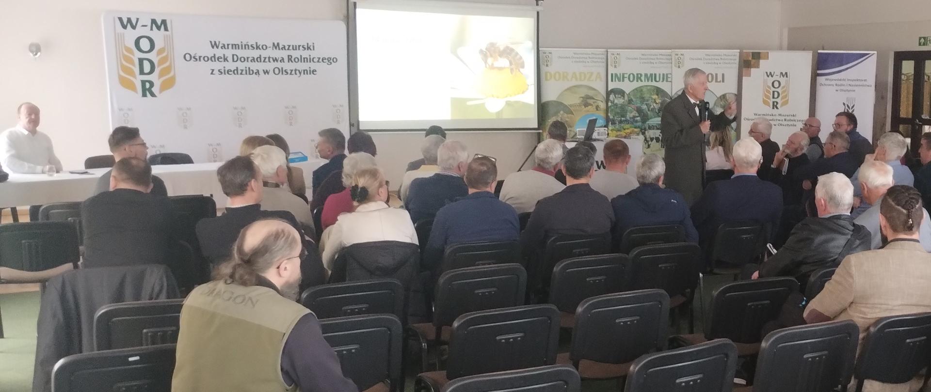 grupa osób siedzi w rzędach naprzeciw stojącego z mikrofonem mężczyzny, za którego plecami wyświetlana jest prezentacja. W prawej części banery firmowe