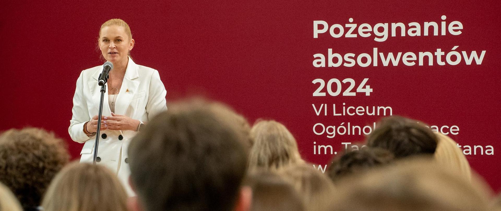 Widok z tyłu na salę. Minister Nowacka stoi przy mikrofonie i przemawia. Za nią na bordowej ścianie napis Pożegnanie absolwentów 2024 VI Liceum Ogólnokształcącego im. Tadeusza Reytana w Warszawie.