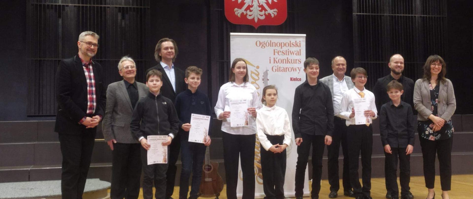 Sala koncertowa Zespołu Szkół Muzycznych w Kielcach, ogromna scena. Na środku sceny stoją laureaci X Ogólnopolskiego Festiwalu i Konkursu "Gitara Viva" w dniu 16 marca 2023 r. Między nimi, po środku stoi uczennica naszej szkoły Lena Torba - najmniejsza ze wszystkich osób, która otrzymała wyróżnienie. Po środku za nimi na ścianie wisi duże godło Polski, pod nim stoi baner z napisem "Gitara Viva!", na suficie wisi mnóstwo żyrandoli. Wśród laureatów stoją jurorzy konkursu, w sumie stoi 13 osób.