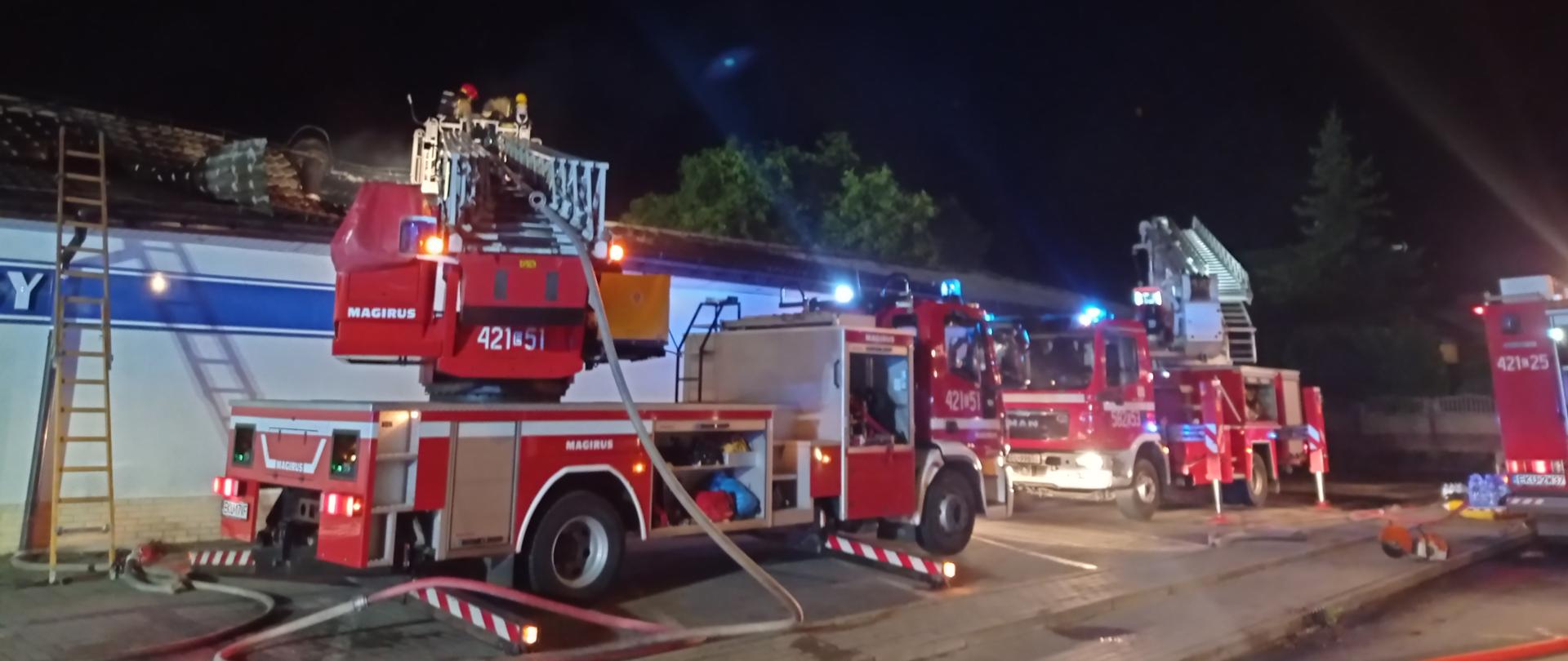  Budynek marketu w porze nocnej. Przed budynkiem marketu trzy samochody specjalne i rozłożone linie gaśnicze. W pojeździe wyposażonym w drabinę mechaniczną strażacy prowadzący akcję gaszenia pożaru.