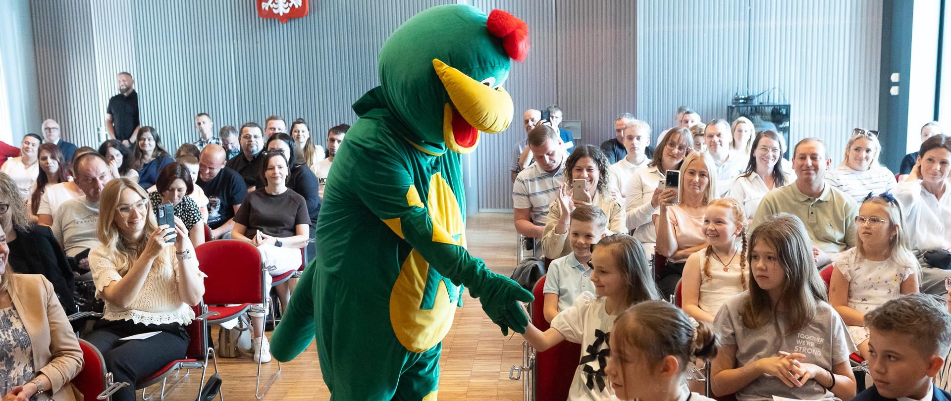 Maskotka - zielony smok przechodzi pomiędzy widownią, na której siedzą dzieci i podaje dzieciom rękę, witając się z nimi.