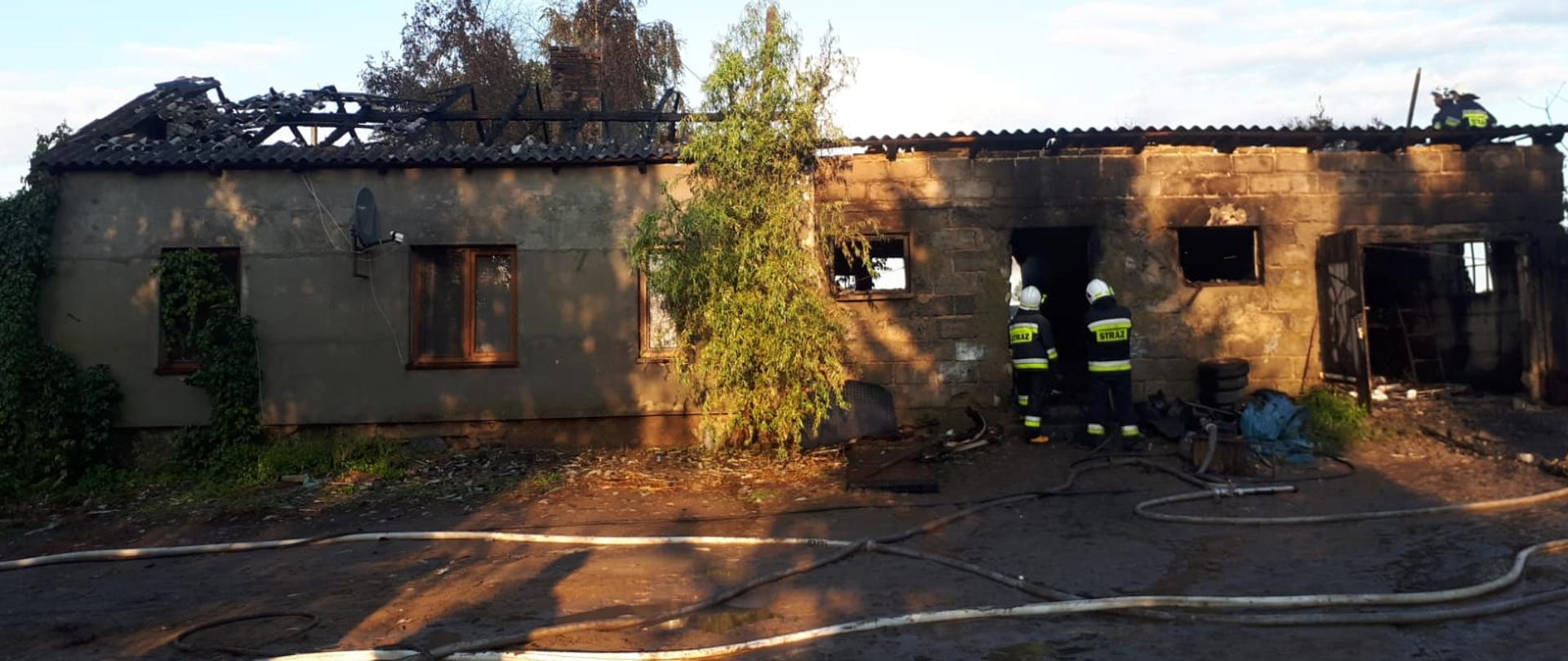 Zdjęcie przedstawia budynek po pożarze. Widać spalony dach oraz pomieszczenia garażowe. Na zdjęciu znajdują się również strażacy. Przed budynkiem widać linie gaśnicze. 