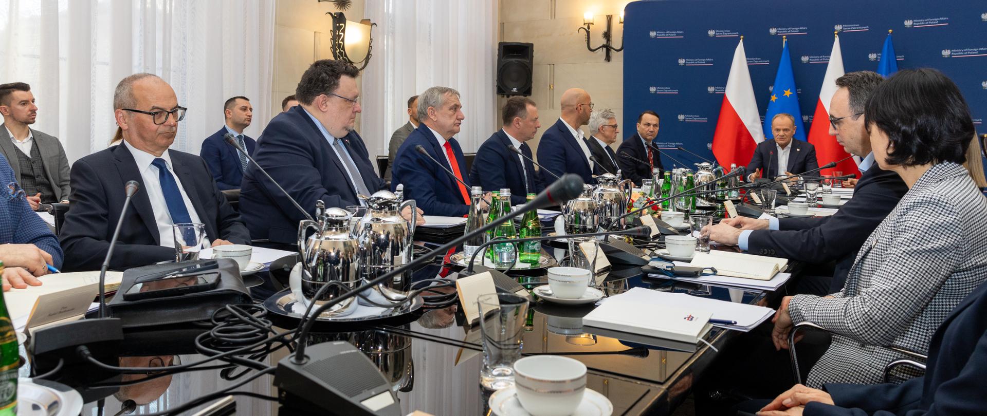 The photo shows a group of people in dark suits. They are sitting at a table with microphones, notebooks, and bottles of water. 