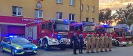 Strażacy oraz policjanci pozują do zdjęcia na tle komendy Państwowej Straży Pożarnej w Wołowie wraz ze swoimi wozami bojowymi i radiowozem z włączonymi sygnałami świetlnymi.
