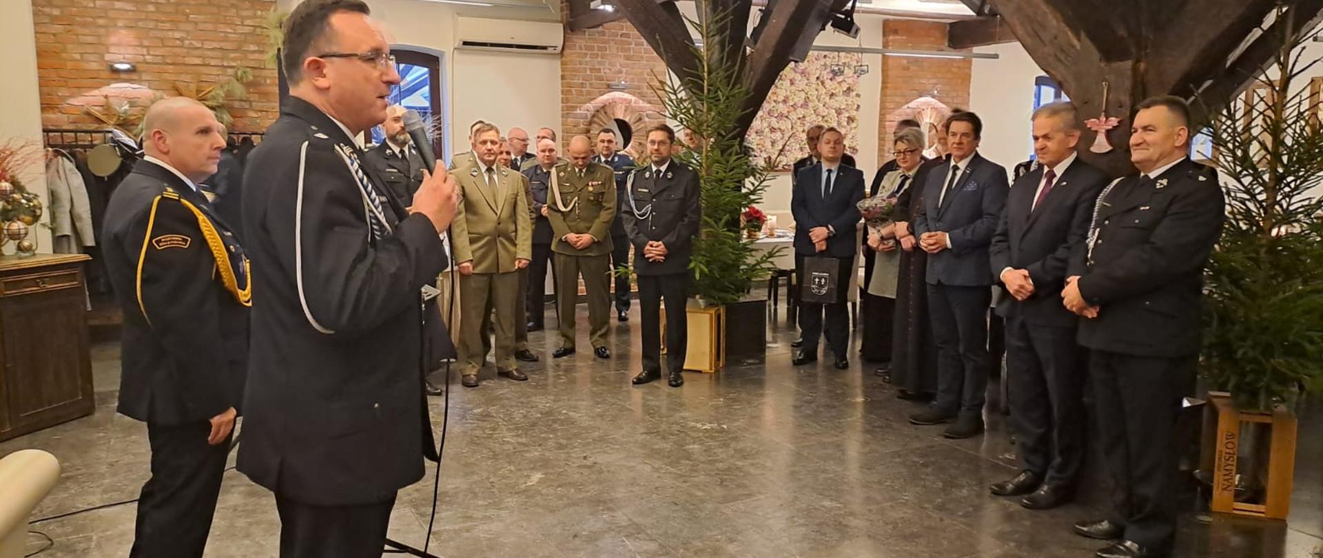 Wnętrze sali bankietowej, z lewej strony przy mikrofonie strażak w mundurze wyjściowym, dalej na wprost i z prawej strony strażacy oraz przedstawiciele samorządów i mundurowi.