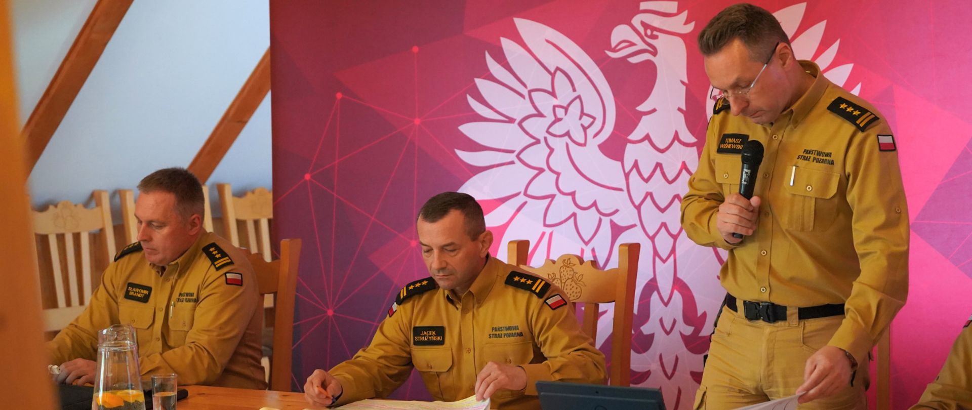 Strażak - komendant wojewódzki PSP - zabiera głos, trzymając mikrofon i mówiąc do komendantów powiatowych, znajduje się na sali, w tle ma ścianę z napisami PSP i orłem rządowym. Obok siedzą kolejni strażacy, za stołem.