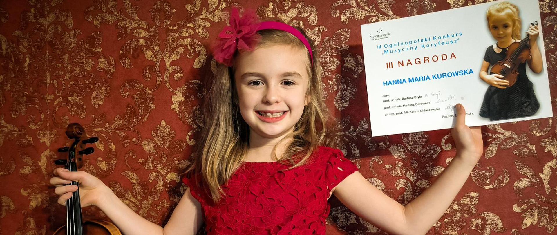 Postać dziewczynki, która w jednej ręce trzyma skrzypce, a w drugiej podniesionej do góry trzyma swój dyplom.