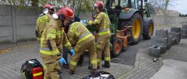 Wypadek podczas prac polowych. Teren jednego z zakładów pracy na terenie Rawicza. Pięciu strażaków na tle ciągnika rolniczego z podczepionym siewnikiem i betonowego płotu udziela kwalifikowanej pierwszej pomocy kobiecie z urazem ręki. Poszkodowana siedzi na krześle obok siewnika podczepionego do ciągnika rolniczego. Jeden ze strażaków podchodzi do torby ze sprzętem do ratownictwa medycznego, stojącej na kostce brukowej.