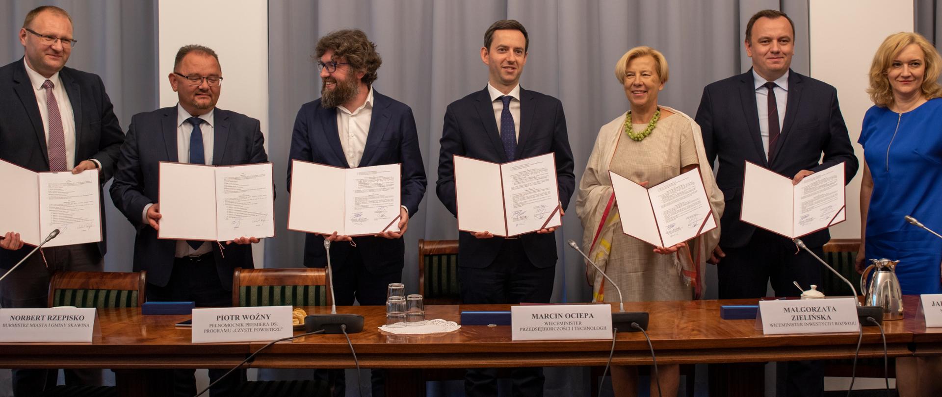 Siedem osób stoi za stołem, sześć z nich trzyma w ręku umowy. 