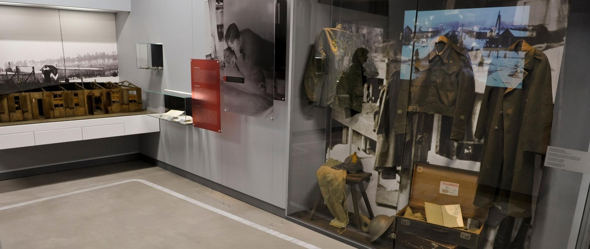 Fragment jednej z wystaw stałych w Muzeum w Łambinowicach. Prawą stronę fotografii zajmuje wysoka szklana gablota, w której znajdują się elementy umundurowania żołnierskiego z odbiciem ekranu z filmem. Z lewej strony drewniana makieta baraku, w którym przetrzymywano jeńców wojennych. Na podłodze duży napis o treści: 1945. Na ścianach powiększone archiwalne fotografie.