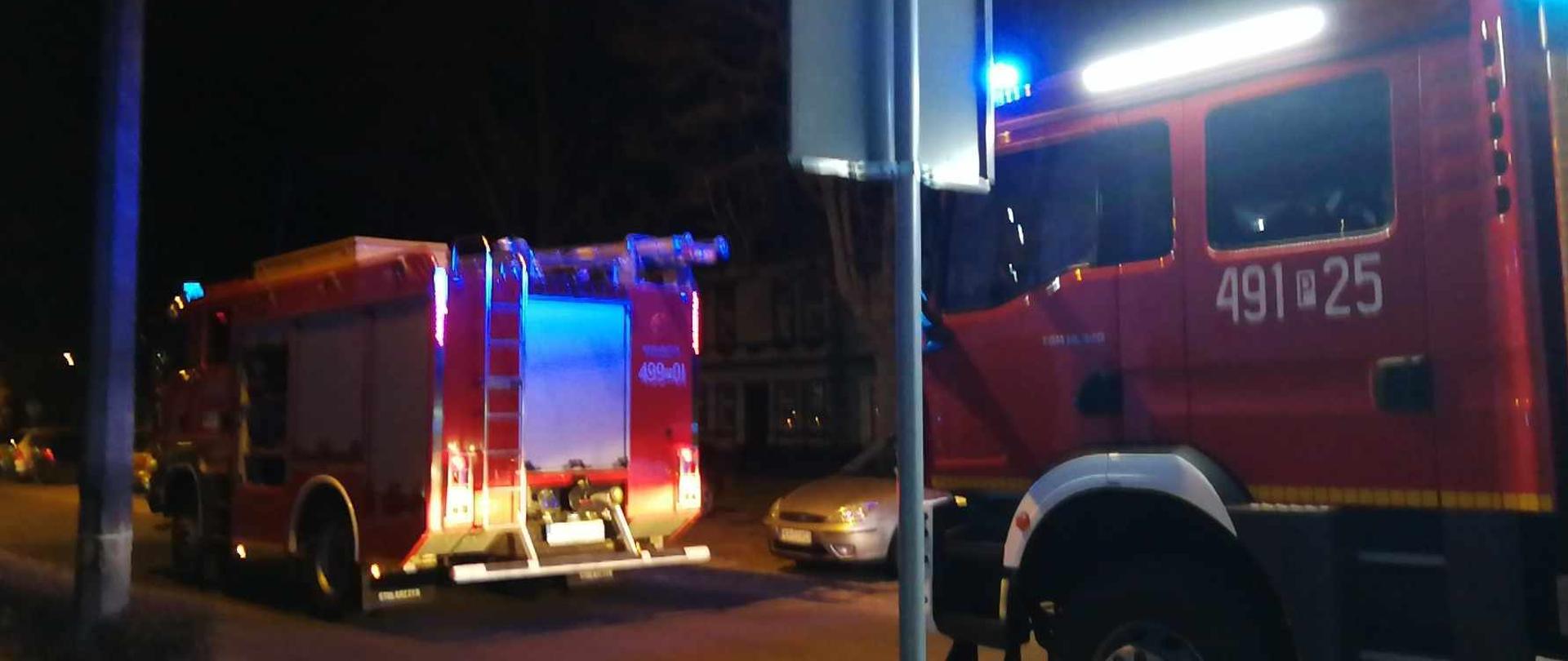 Dwa pojazdy pożarnicze stoją na drodze przed budynkiem mieszkalnym. Pojazdy mają włączone sygnały błyskowe. Trwają działania związane z likwidacją zagrożenia z udziałem tlenku węgla.