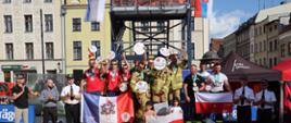 Na podium zawodnicy zawodów z flagami, w ich otoczeniu strażacy i osoby cywilne