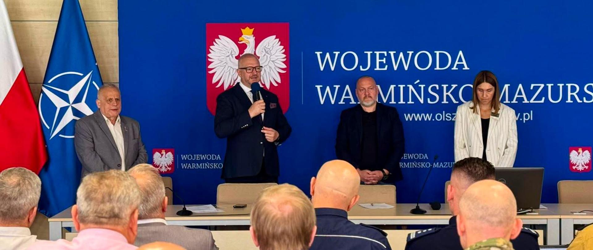 Cztery osoby w oficjalnych strojach pozują do zdjęcia na tle ścianki Wojewody Warmińsko-Mazurskiego. Wśród nich znajdują się wojewoda, wicewojewoda, pierwszy zastępca dyrektora Wydziału Bezpieczeństwa i Zarządzania Kryzysowego oraz kierownik jednego z oddziałów tego wydziału.