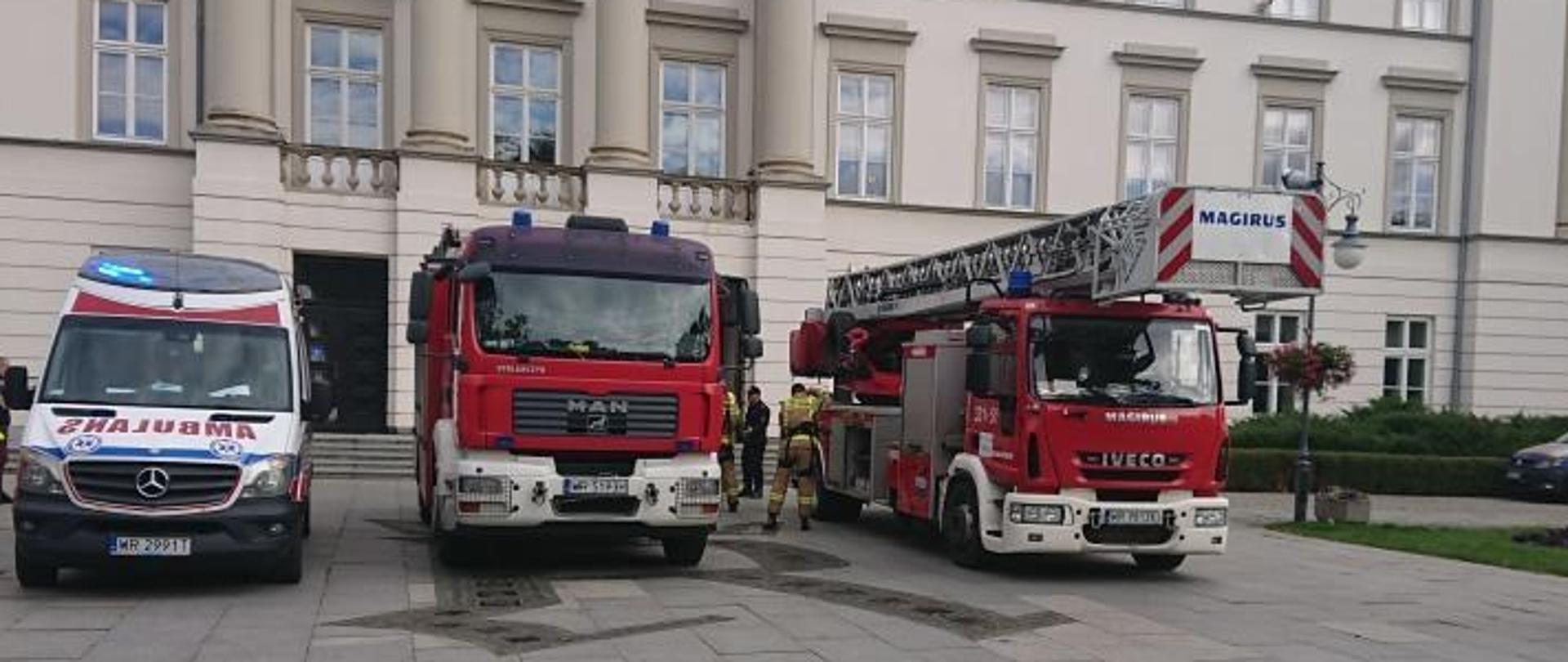 Zdjęcie wykonane przed Urzędem Miejskim w Radomiu podczas próbnej ewakuacji pracowników oraz interesantów. Na zdjęciu widać ustawione od lewej: karetkę pogotowia ratunkowego oraz dwa pojazdy pożarnicze Państwowej Straży Pożarnej. Karetka w kolorze białym z czerwonymi i niebieskimi elementami zabudowy. Pojazdy pożarnicze w kolorze czerwonym z białymi i srebrnymi elementami zabudowy. Jeden z pojazdów to pojazd ratowniczo – gaśniczy natomiast drugi to autodrabina mechaniczna. 