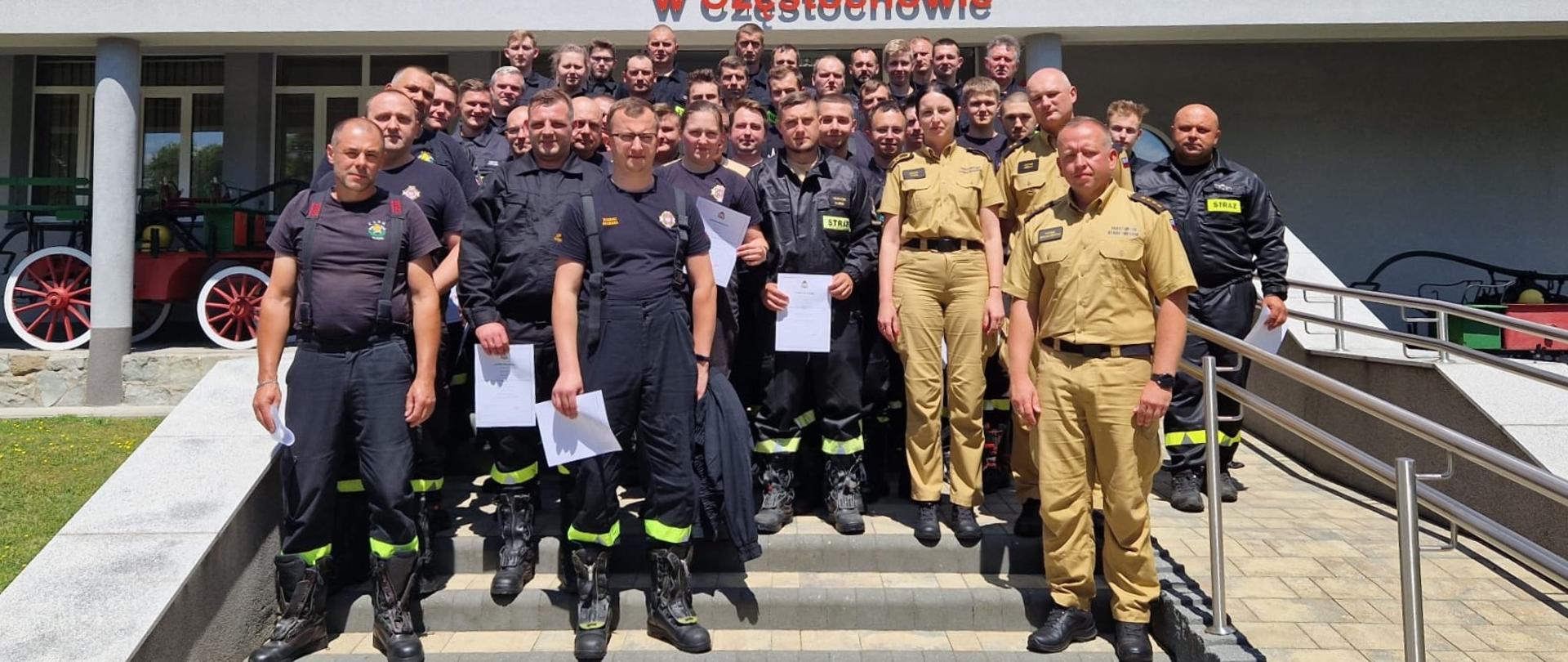 Zdjęcie grupowe uczestników szkolenia wraz z komisją egzaminacyjną stojących na schodach przed wejściem głównym do budynku Komendy Miejskiej Państwowej Straży Pożarnej