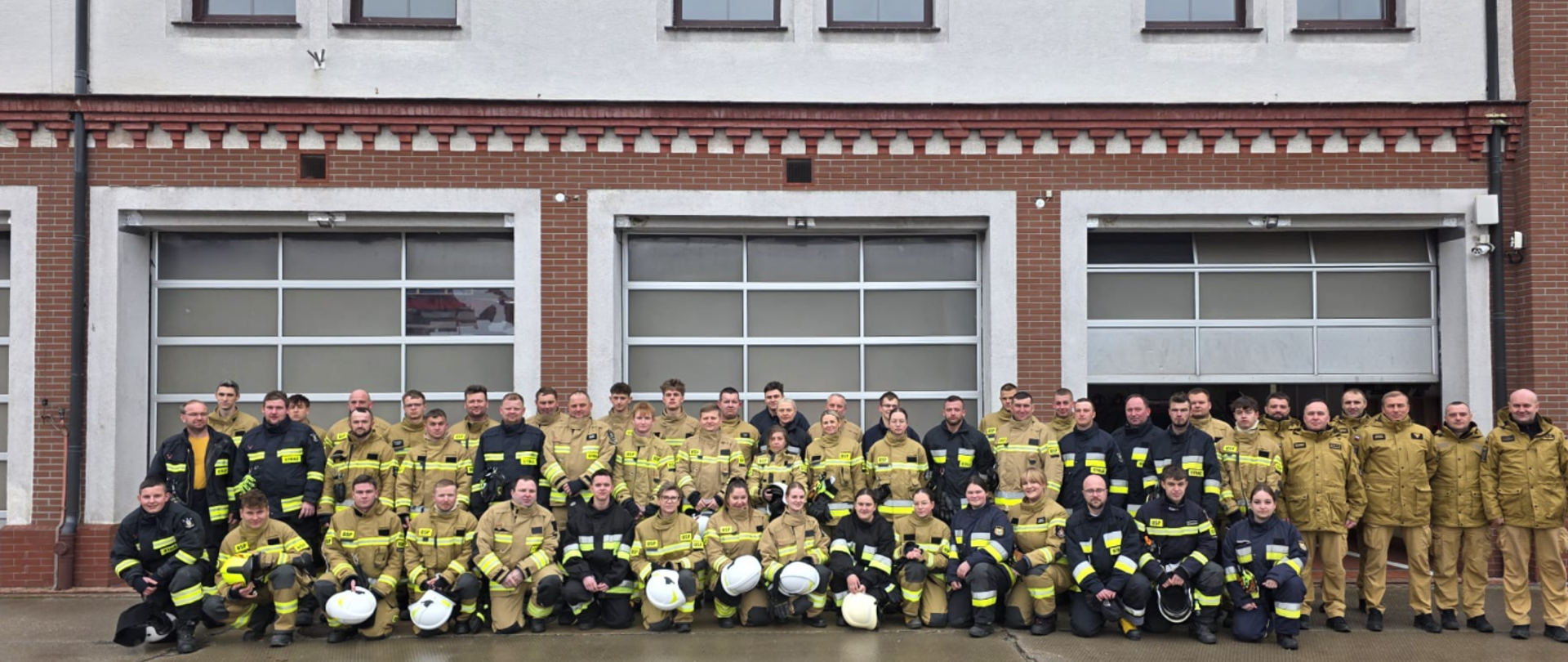 Zdjęcie grupowe dużej jednostki straży pożarnej na tle nowoczesnej remizy. Kilkudziesięciu strażaków i strażaczek ustawionych w trzech rzędach. Wszyscy są ubrani w profesjonalne mundury specjalne – przeważają stroje w kolorze piaskowym, część osób ma na sobie mundury czarne.